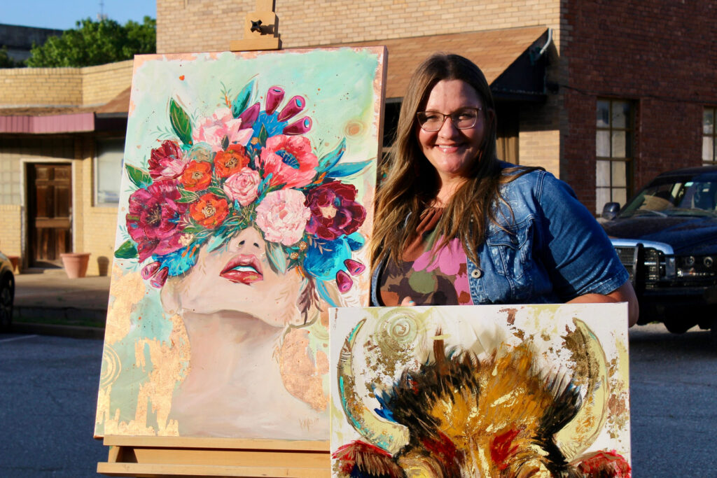Woman smiling while holding and standing beside two colorful paintings, one of a flower-faced woman and another abstract piece, outdoors.