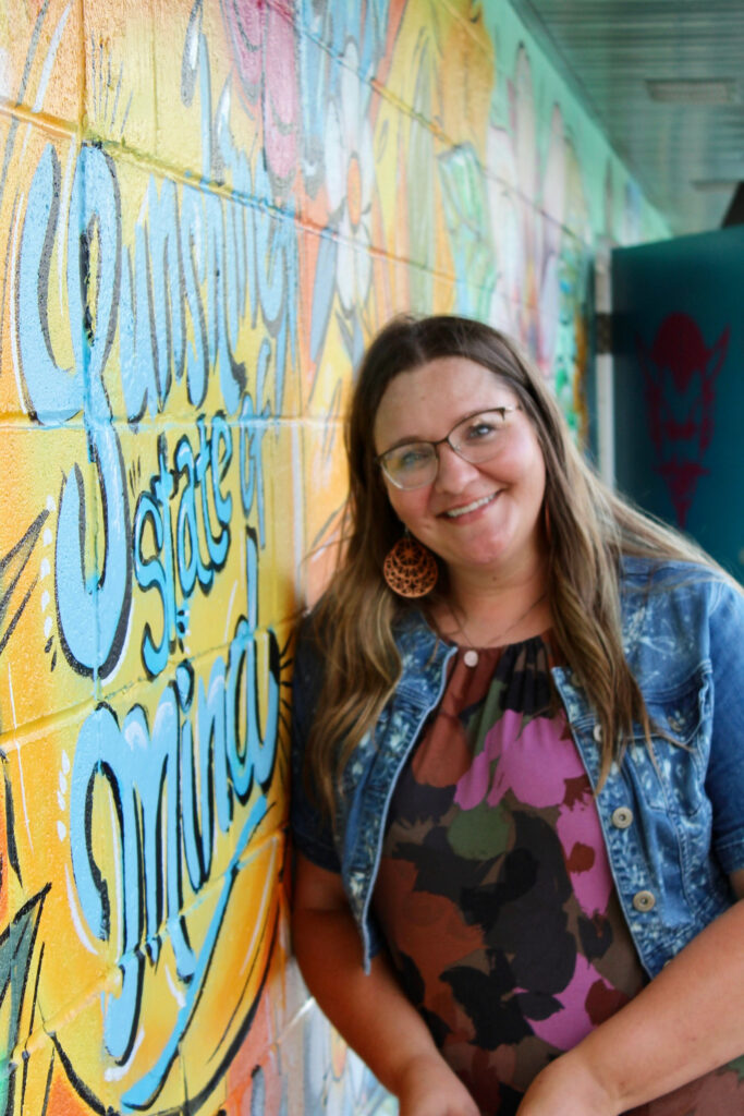 Woman standing in front of a vibrant mural on a brick wall that says “Sunshine state of mind,” surrounded by large floral artwork.